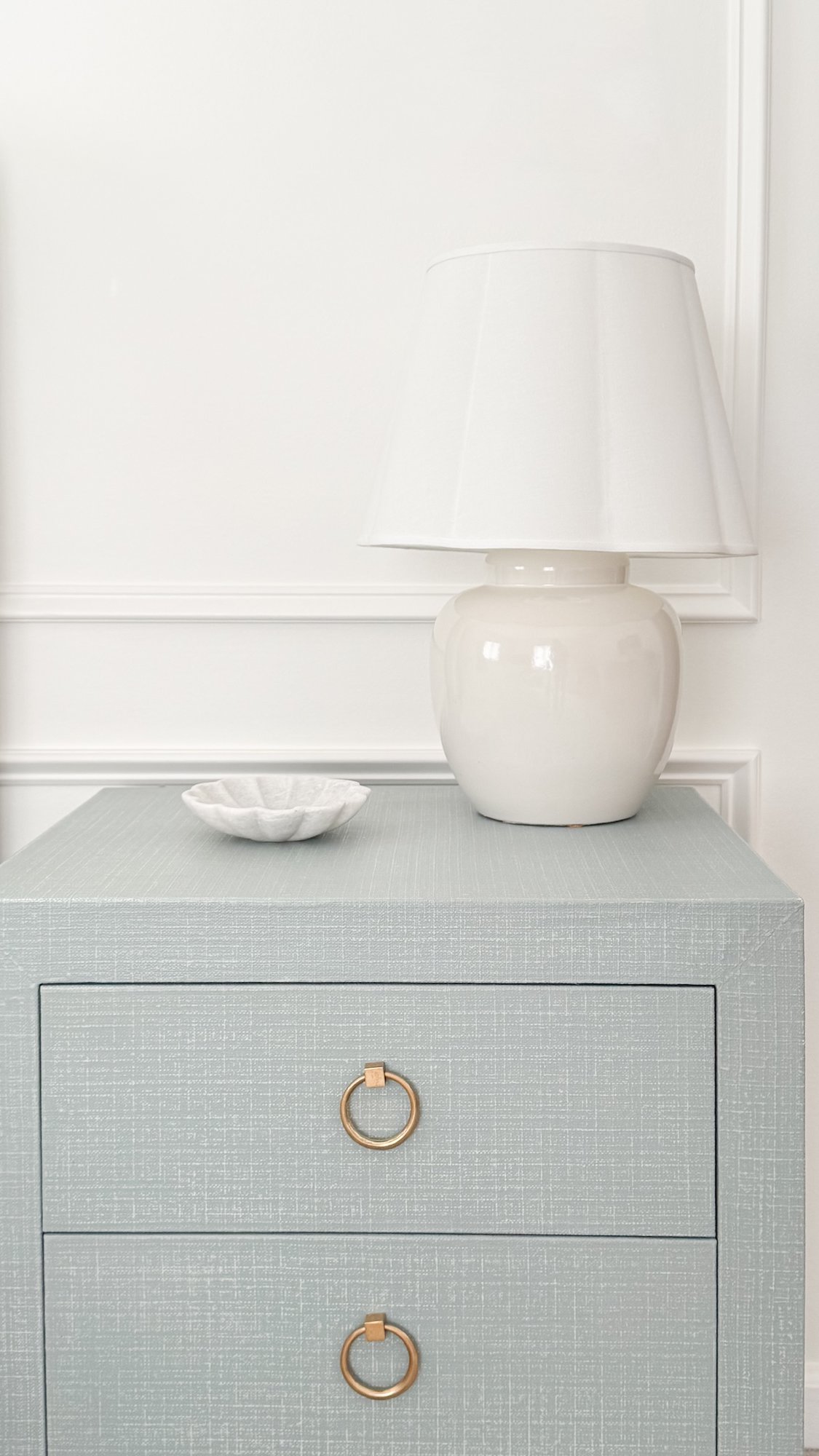 A light blue textured nightstand with gold ring pulls holds a white ceramic lamp and a small white dish, against a white paneled wall—a stylish example for those learning how to measure furniture for the perfect fit.