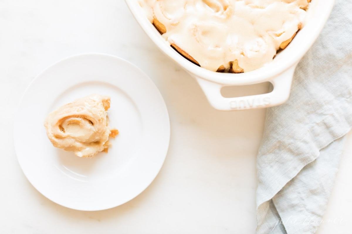 A white oval pan full of frosted caramel rolls, with one on a small serving plate to the side.