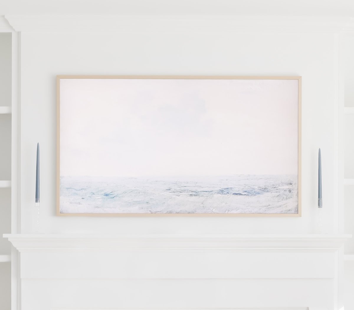 A framed minimalist seascape, displayed as Samsung Frame TV Art, hangs above a white mantel, flanked by two tall blue taper candles in glass holders.