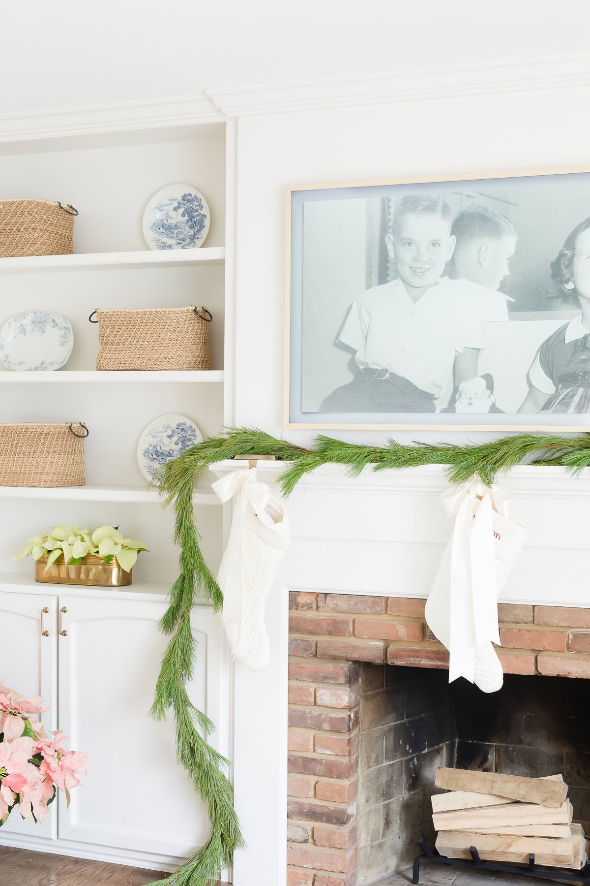 A white fireplace decorated with a pine garland and two white stockings, with Samsung Frame TV Art displaying a vintage family photo above and woven baskets and blue plates on nearby shelves.