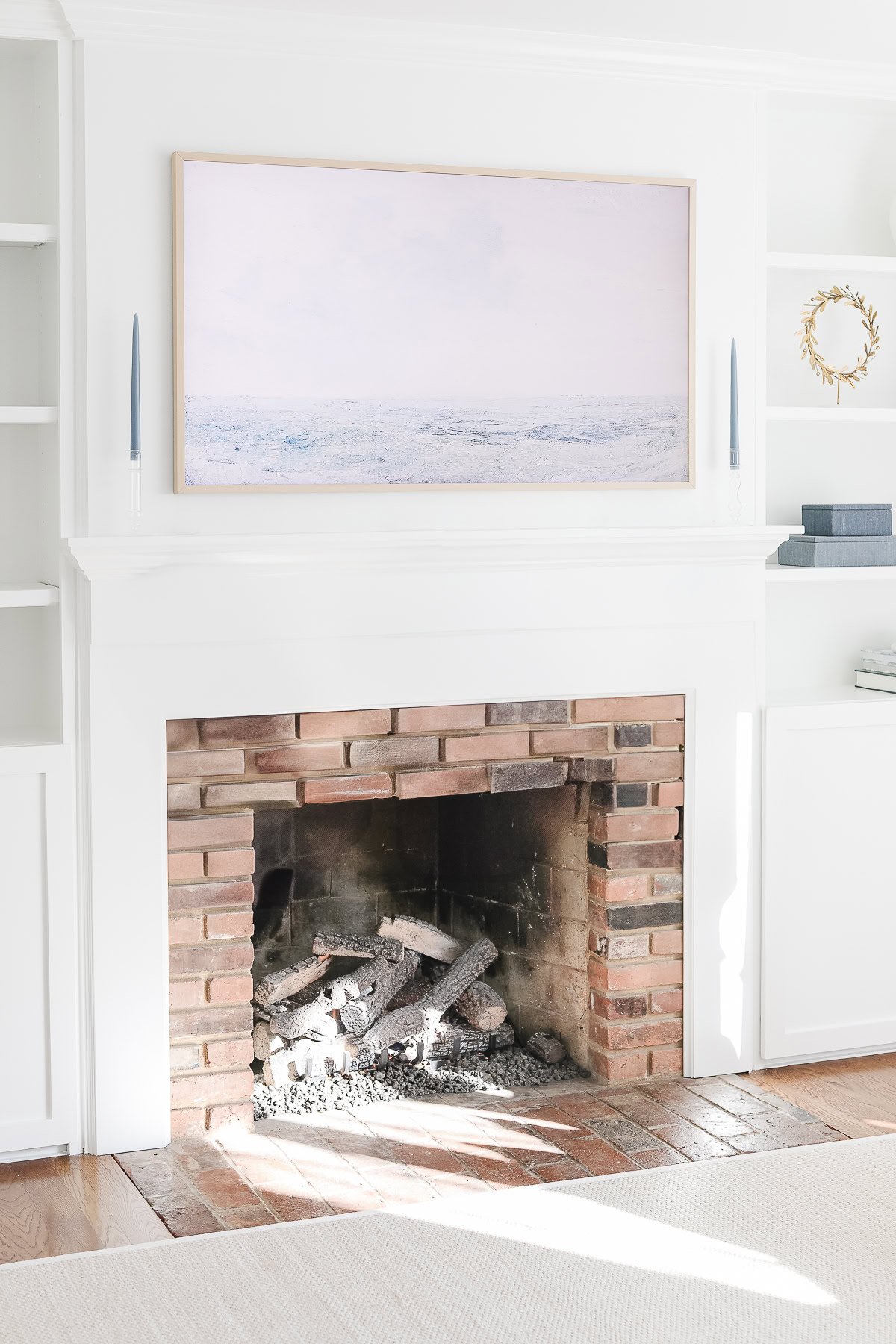 A white fireplace with a brick interior, two tapered candles, and a Samsung Frame TV Art displaying a seascape above, flanked by built-in white shelves.