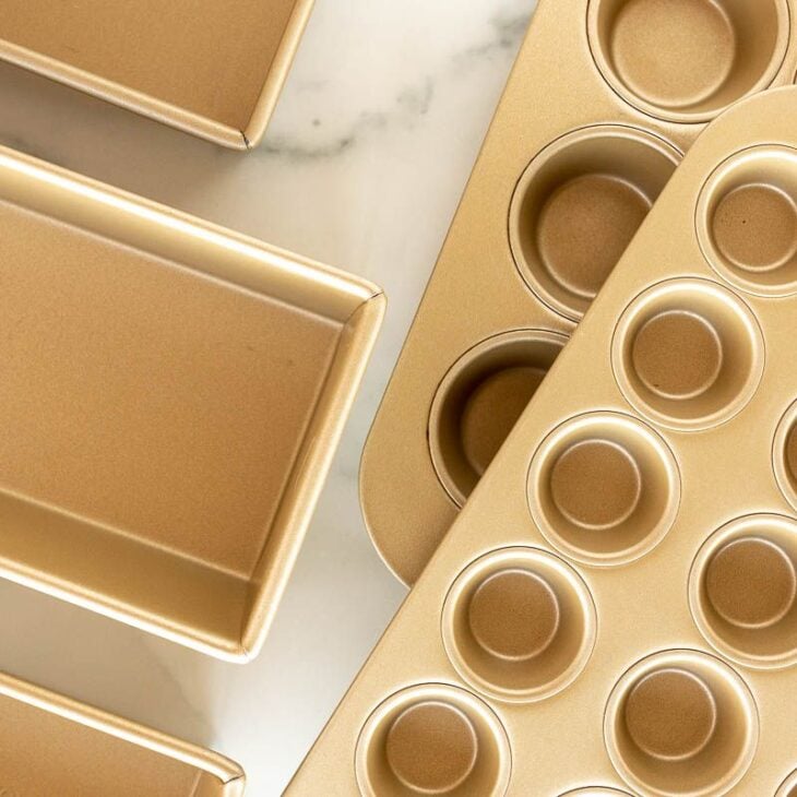 Una variedad de moldes dorados para pasteles, moldes para pan y tamaños de moldes para muffins colocados sobre una superficie de mármol.