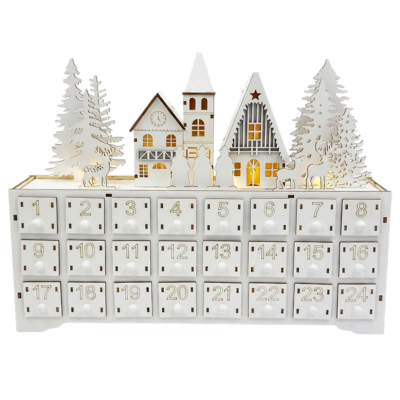 A white wooden advent calendar featuring 24 numbered drawers, topped with a decorative scene of illuminated houses, evergreen trees, and deer.