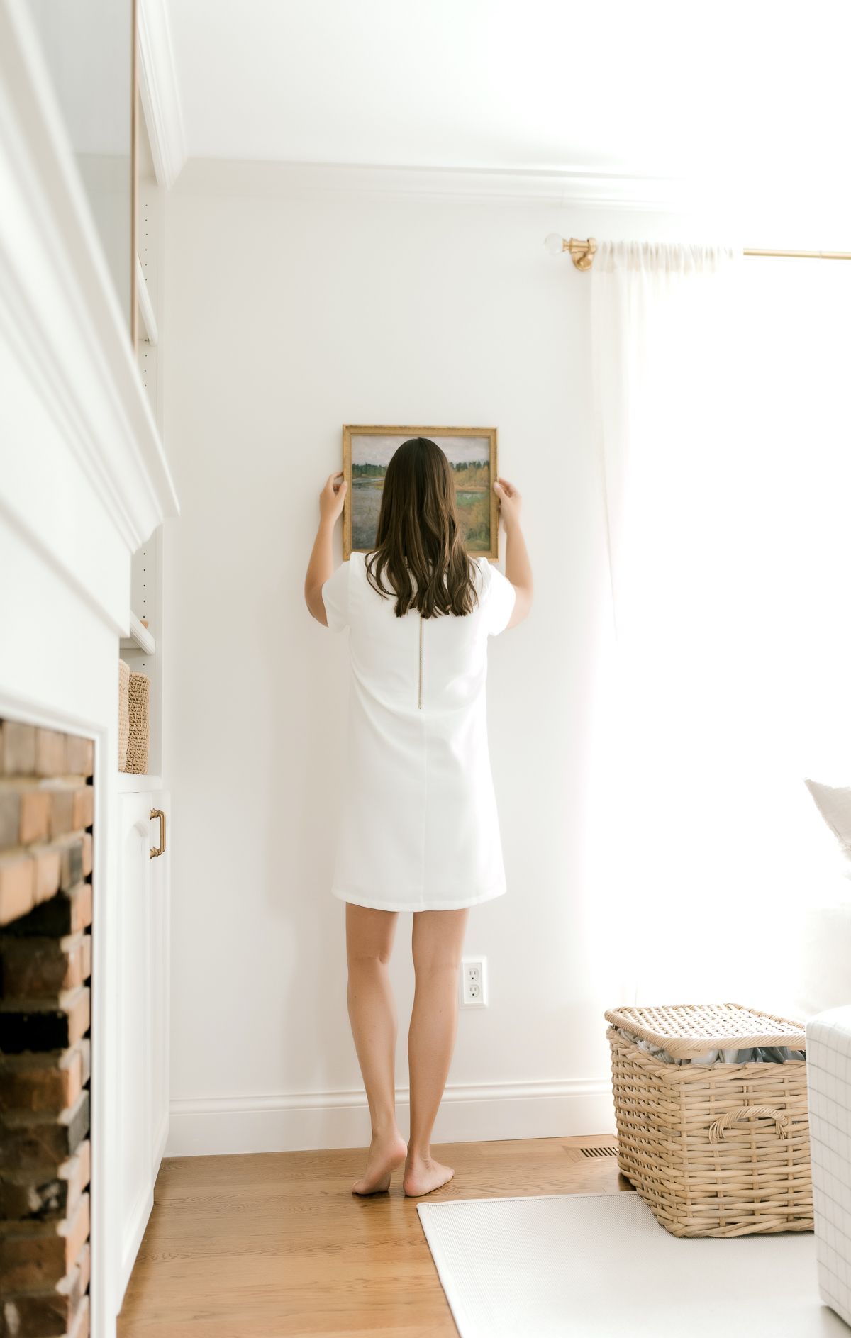 A woman in a white dress hanging a painting in a living room
