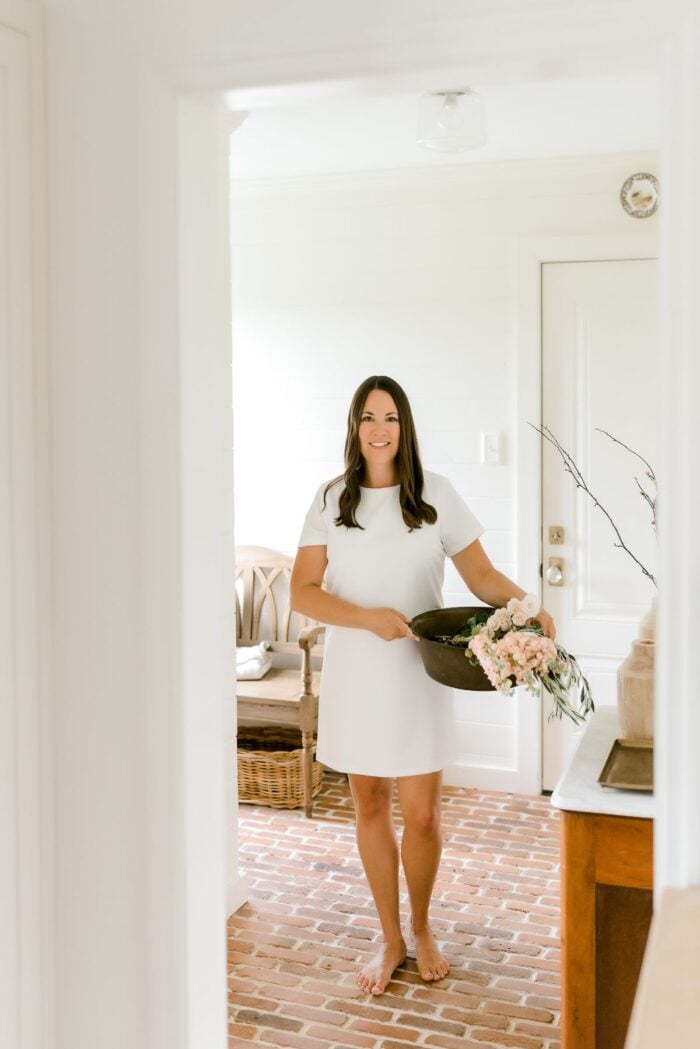 A woman in a white dress, carrying a container of fresh flowers in a white room
