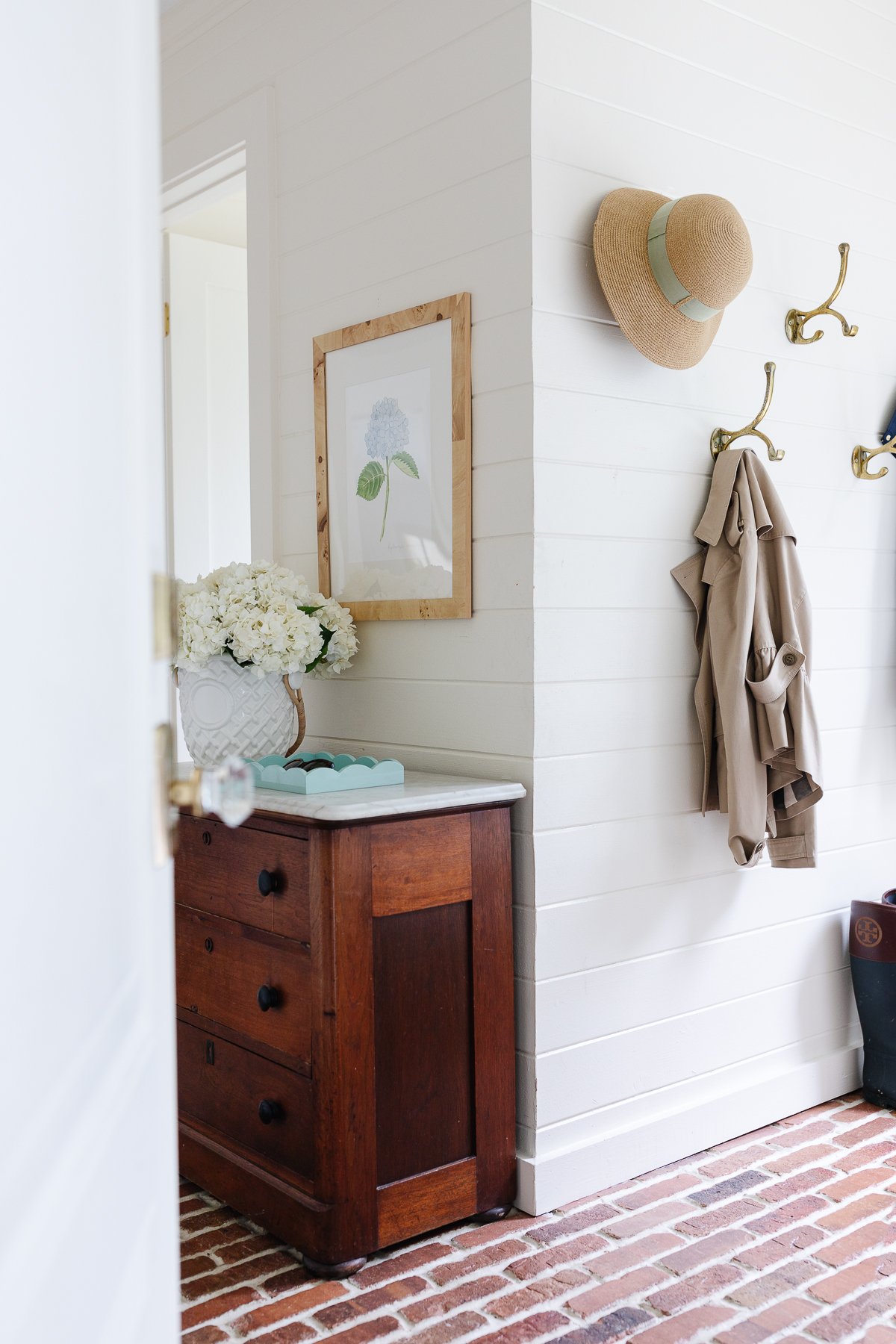 white paneled walls in a mud room with brick floors and brass hooks.