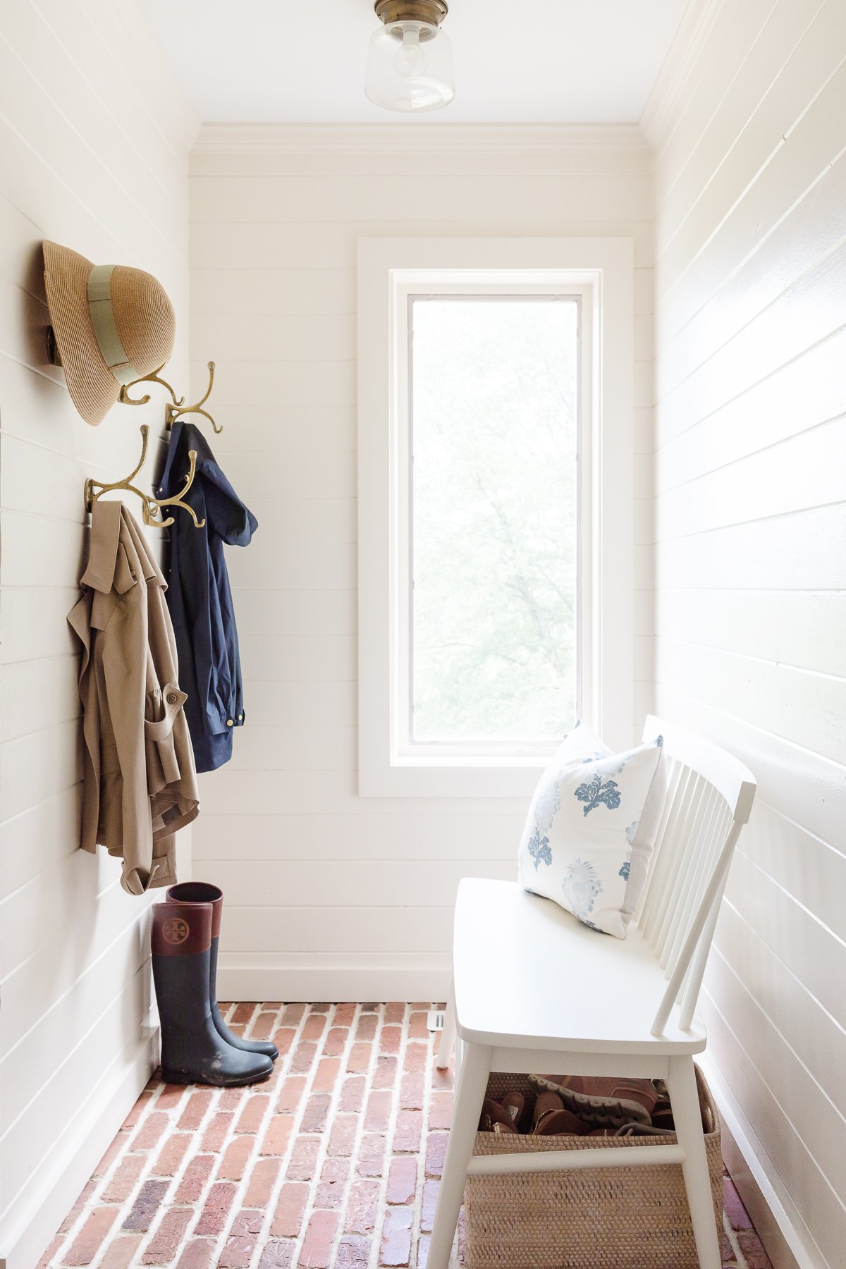 white paneled walls in a mud room with brick floors and brass hooks.