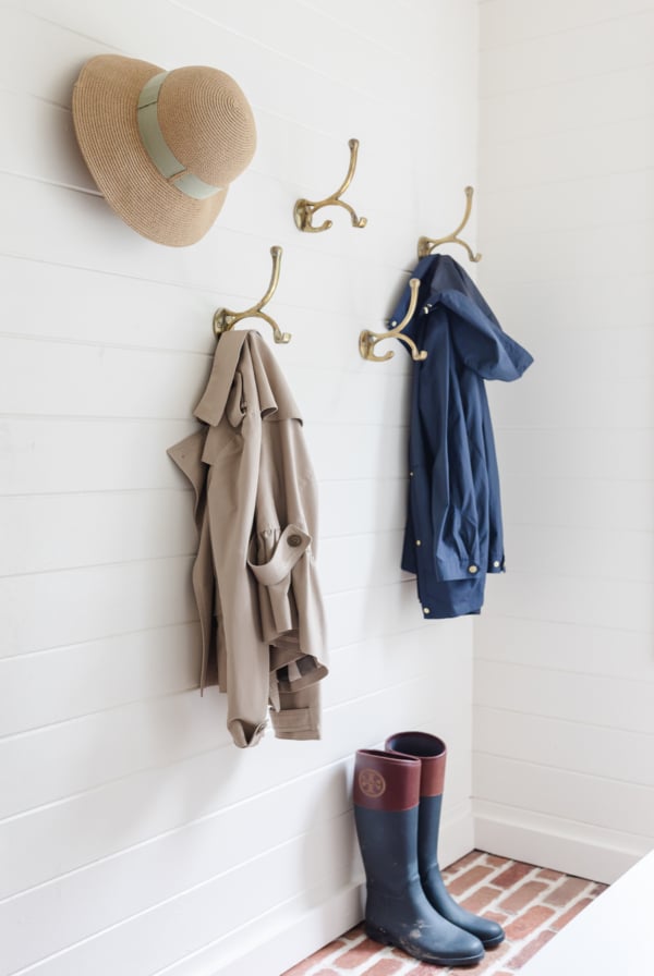 A white paneled mudroom with brass hooks on the wall holding hats and jackets.