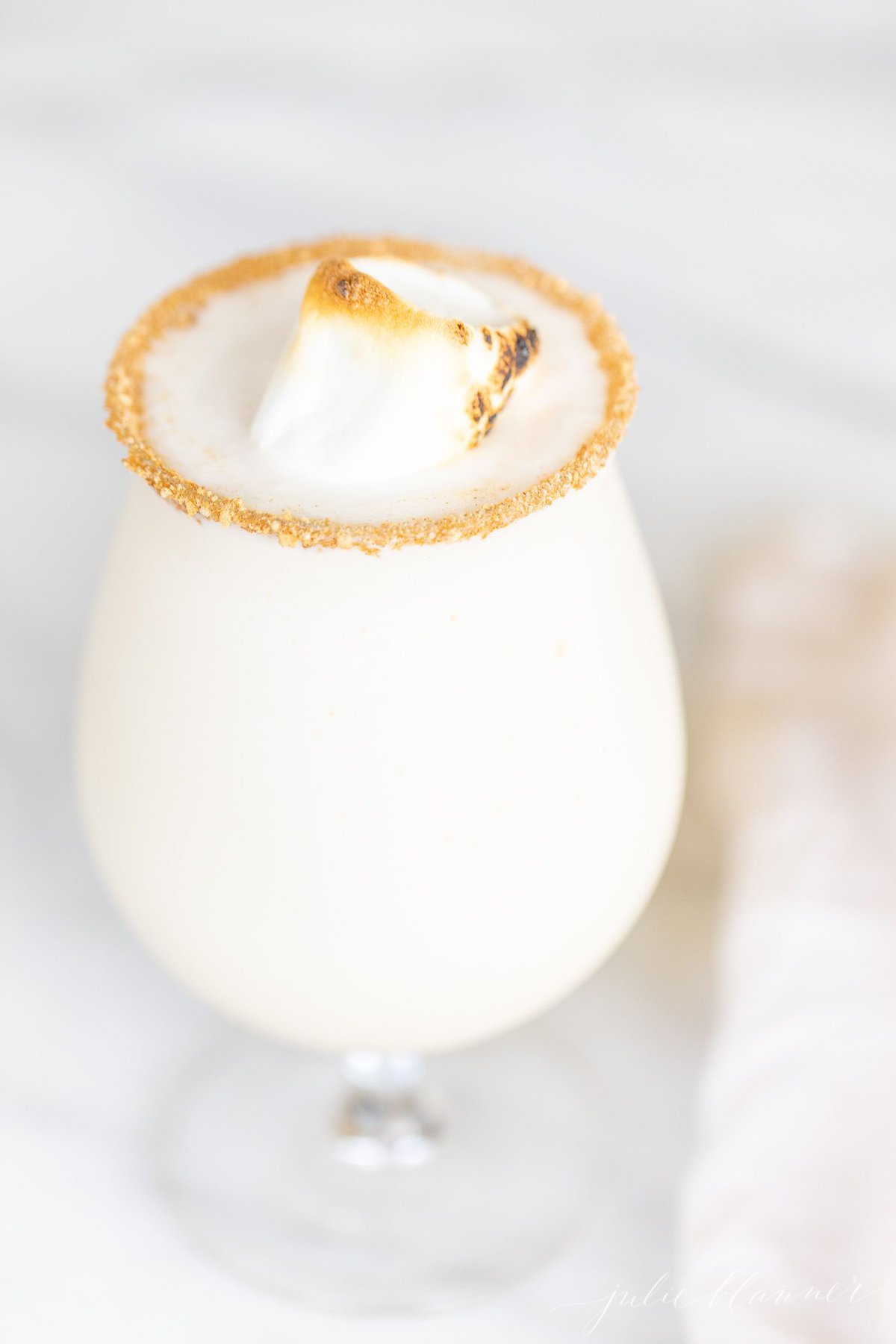 A clear glass with a creamy white Christmas cocktail, topped with a toasted marshmallow.