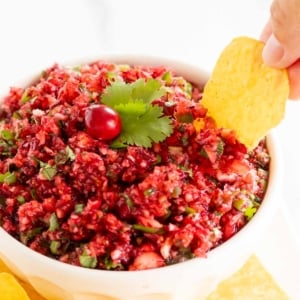 A hand dipping a tortilla chip into a bowl of fresh Christmas cranberry salsa.