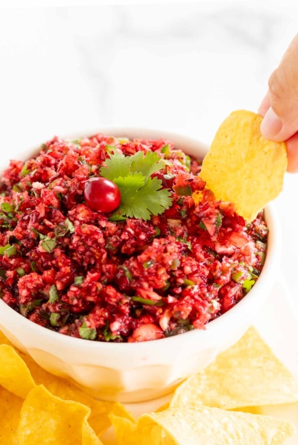 A hand dipping a tortilla chip into a bowl of fresh Christmas cranberry salsa.