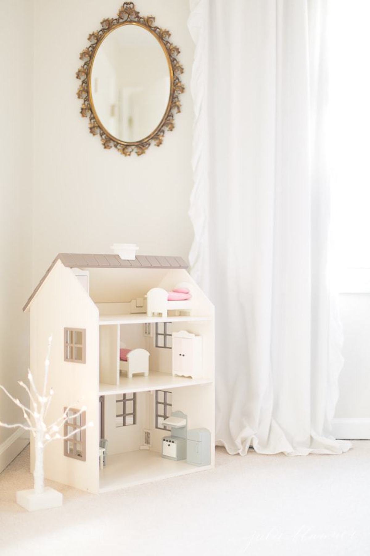 A doll house in a white room with a fairy light tree.