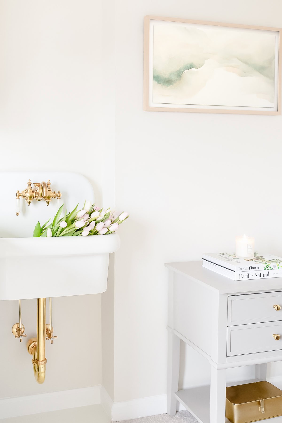 A white wall-mounted sink with gold fixtures holds a bouquet of white tulips; beside it is a gray table with books, a candle, and Samsung Frame TV Art displayed above as striking abstract artwork.