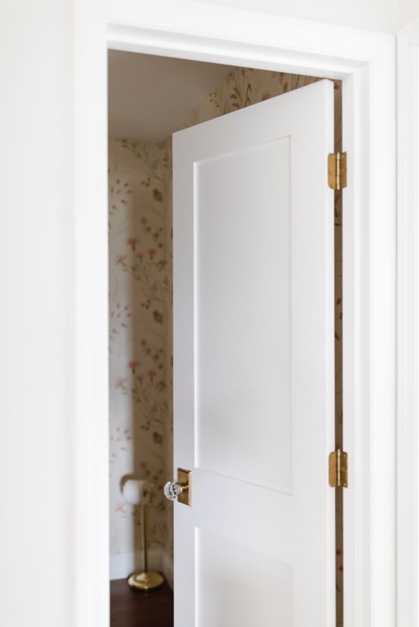 A partially open white door revealing a glimpse of a bathroom with floral wallpaper.