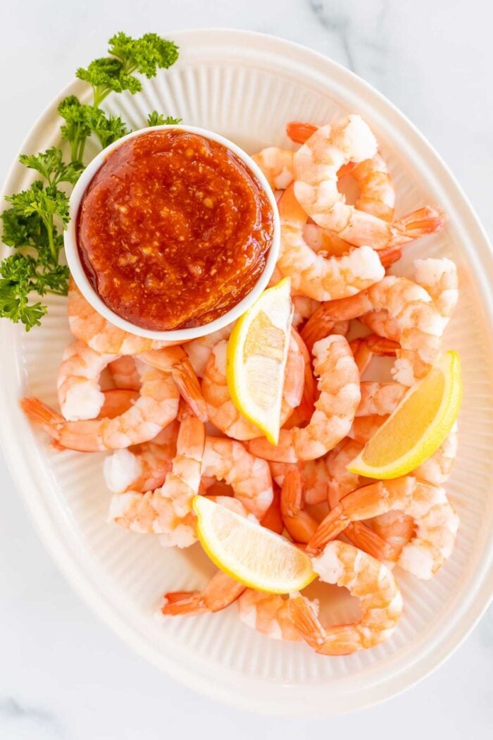 An oval platter of roasted shrimp cocktail with a homemade cocktail sauce in a white bowl.