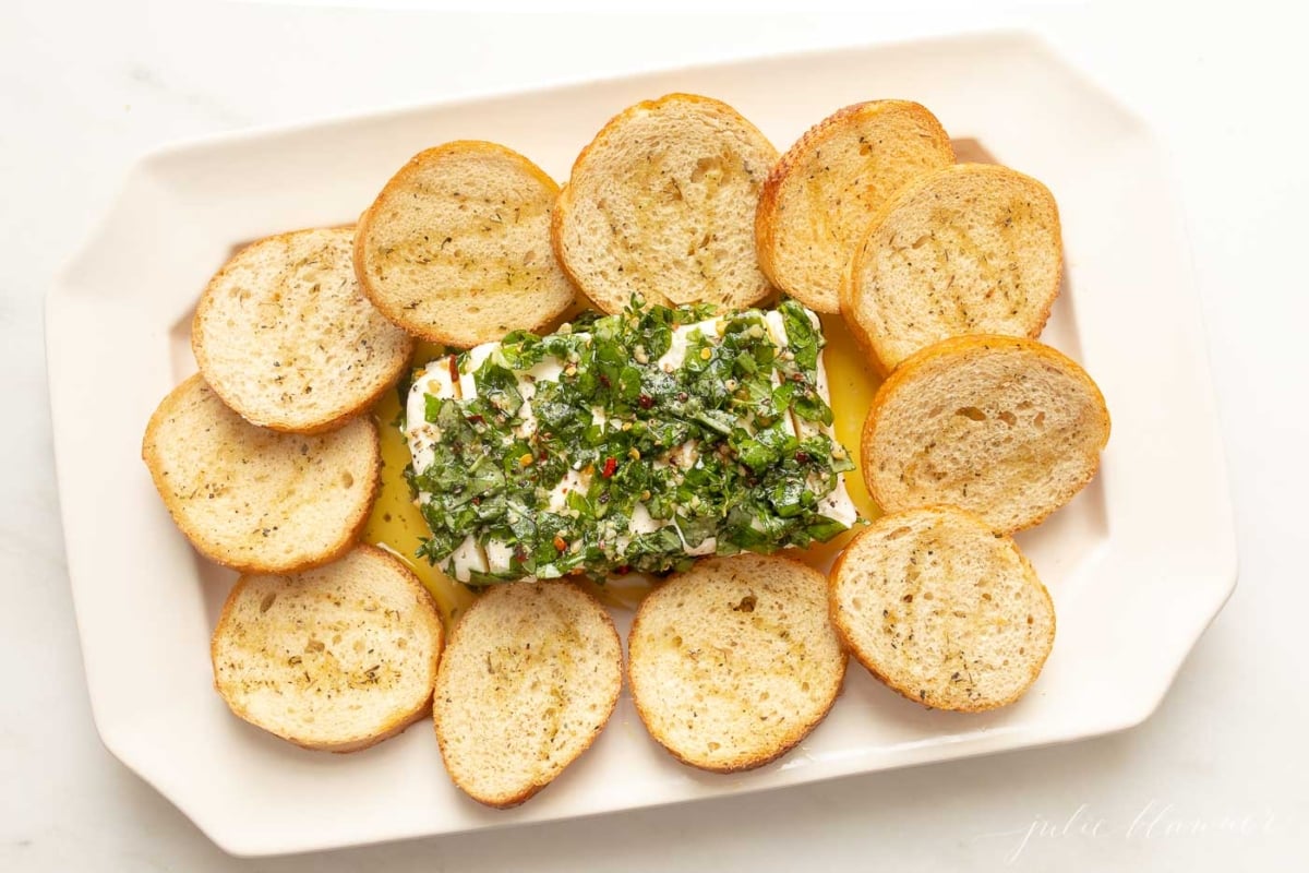 Marinated cheese surrounded by crostini on a white platter.