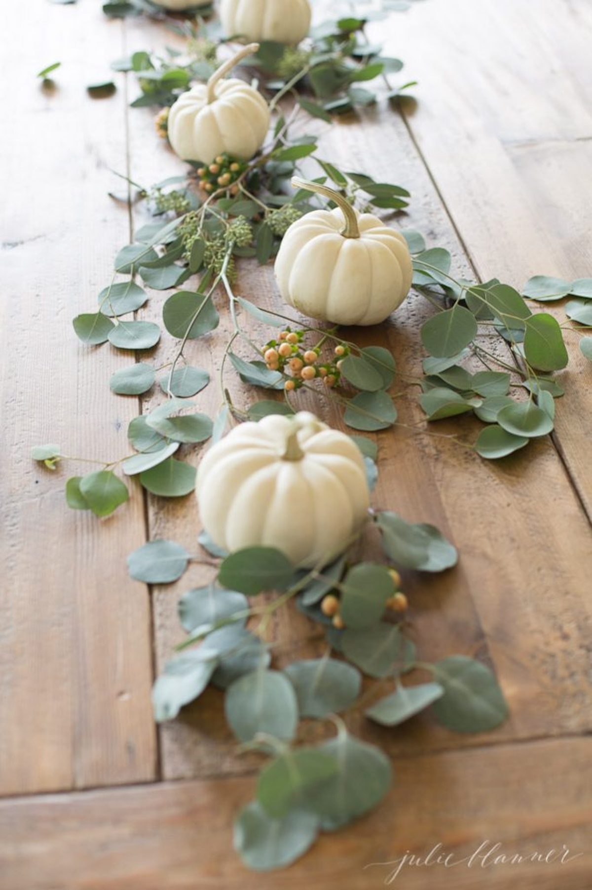 Thanksgiving table decor featuring white pumpkins and eucalyptus leaves on a wooden table.