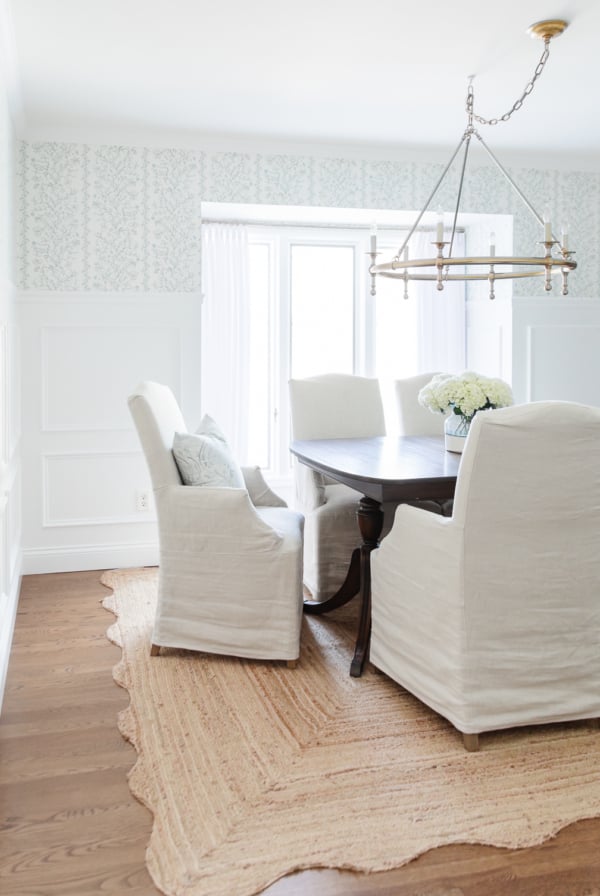 Elegant dining room featuring a dark wood table, white slipcovered chairs, a beige rug, and a gold chandelier, with Benjamin Moore Chantilly Lace walls and floral wallpaper.