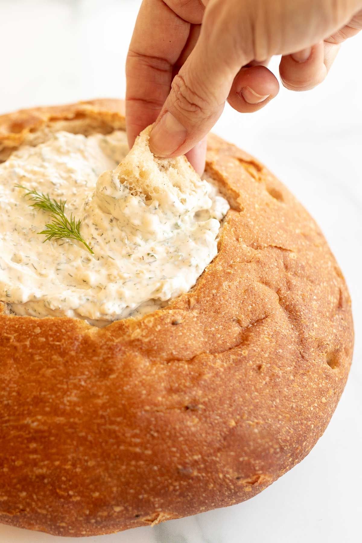 A hand dipping into a bread bowl full of dill dip