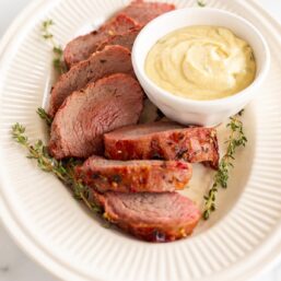 A grilled beef tenderloin on an oval platter with a bowl of dijon mustard sauce.