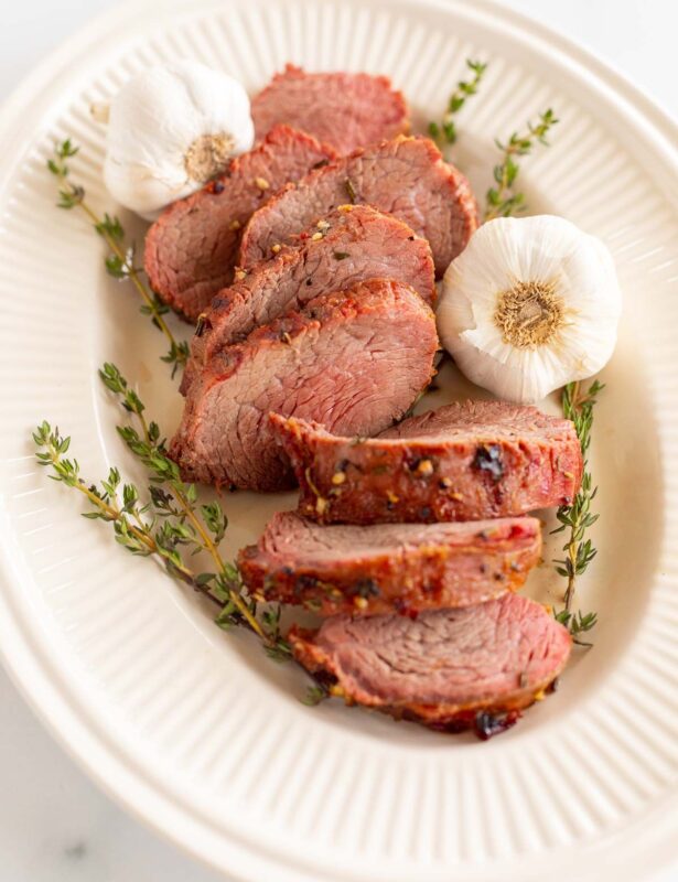 A platter of sliced beef tenderloin, garnished with fresh thyme and garlic.