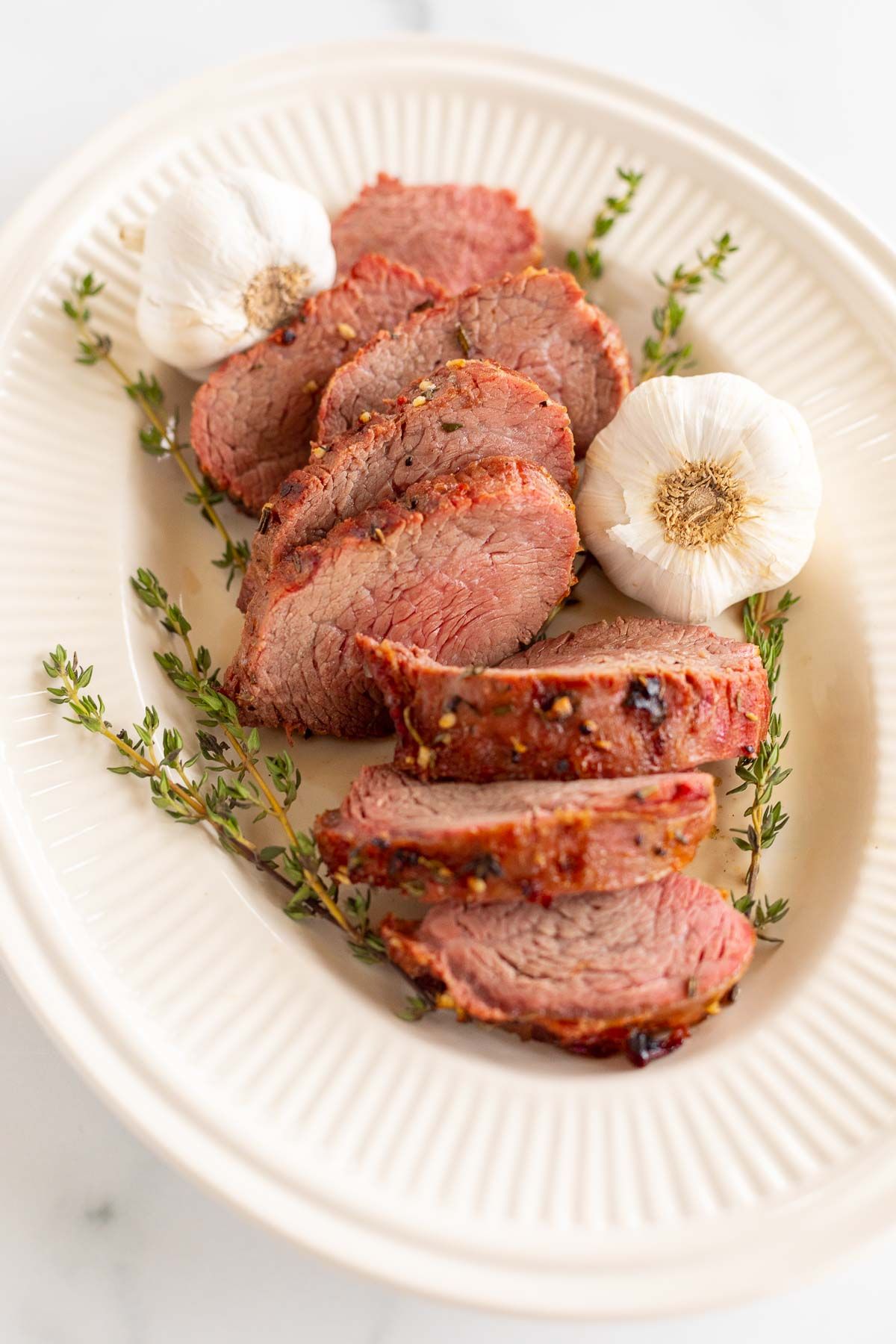A platter of sliced beef tenderloin, garnished with fresh thyme and garlic.