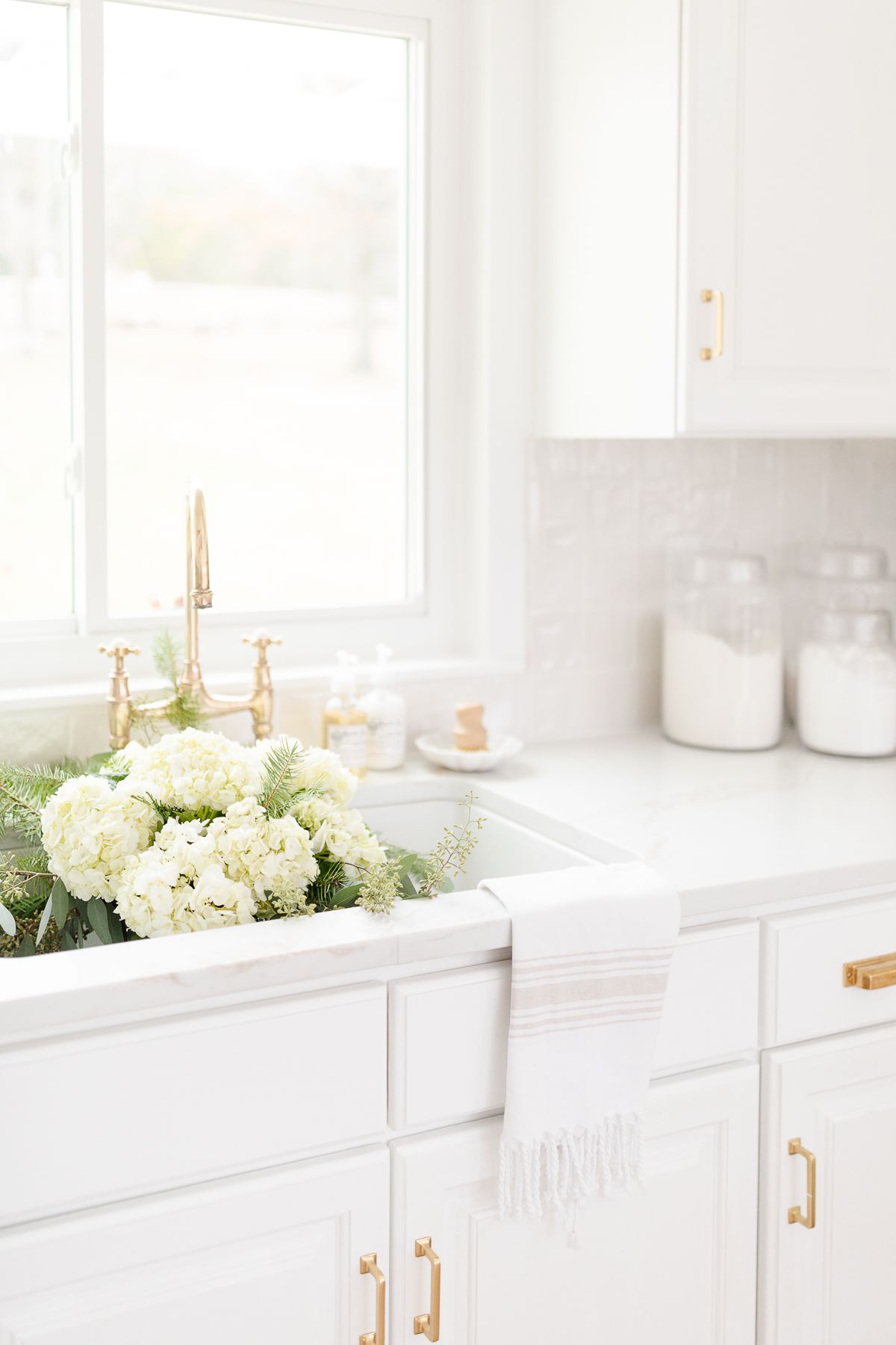 A white kitchen with flower in the sink, brass faucet and white backsplash