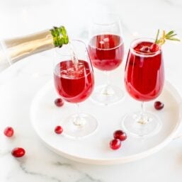 A white tray with 3 glasses full of poinsettia cocktail, champagne being poured in