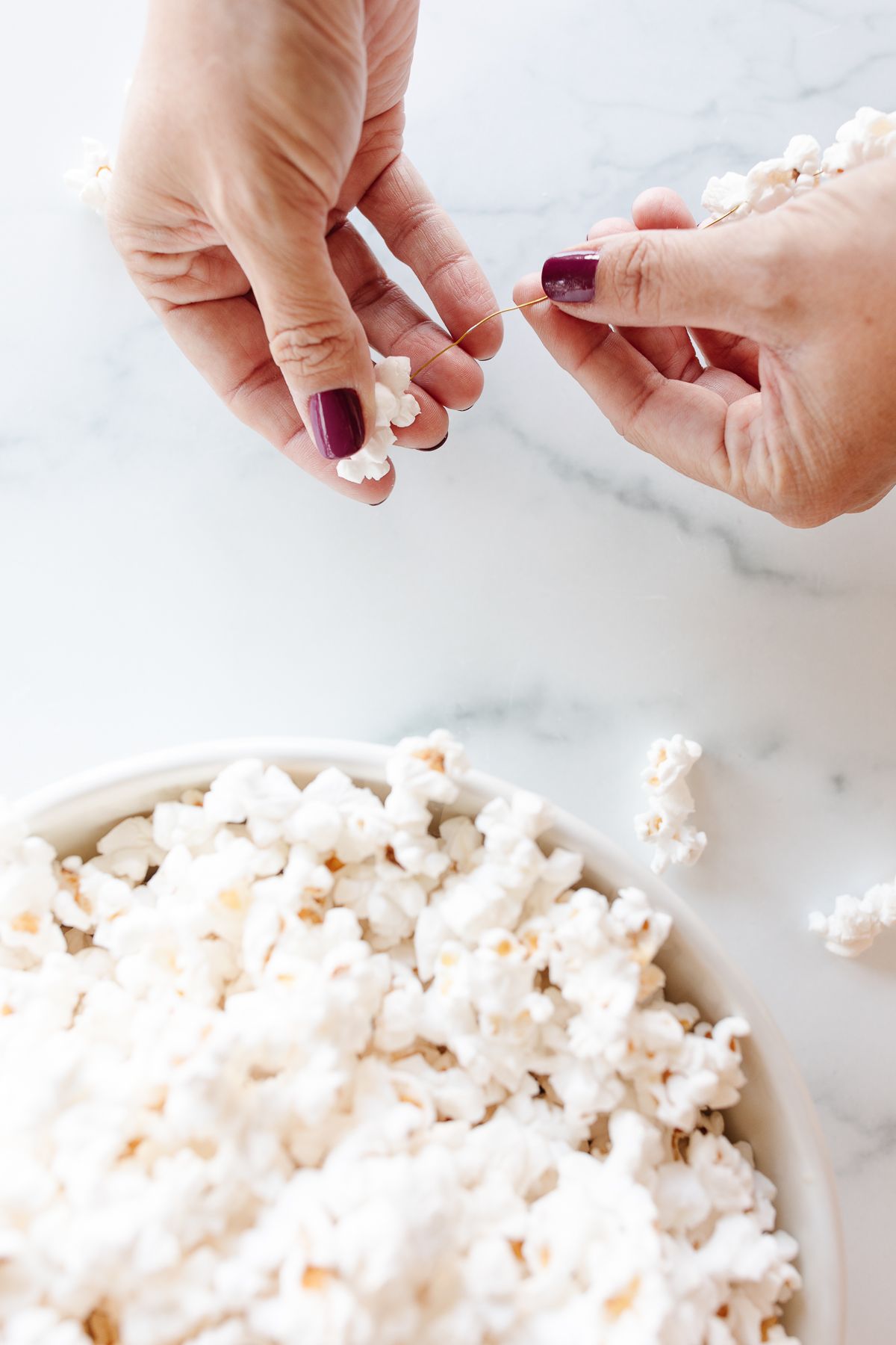 A bowl of popcorn and a woman's hands demonstrating how to make a garland of popcorn.