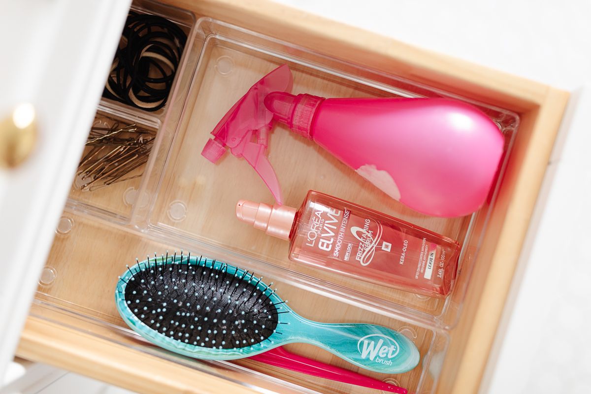 A clear plastic bathroom drawer organizer with a spray bottle, hair brushes and accessories