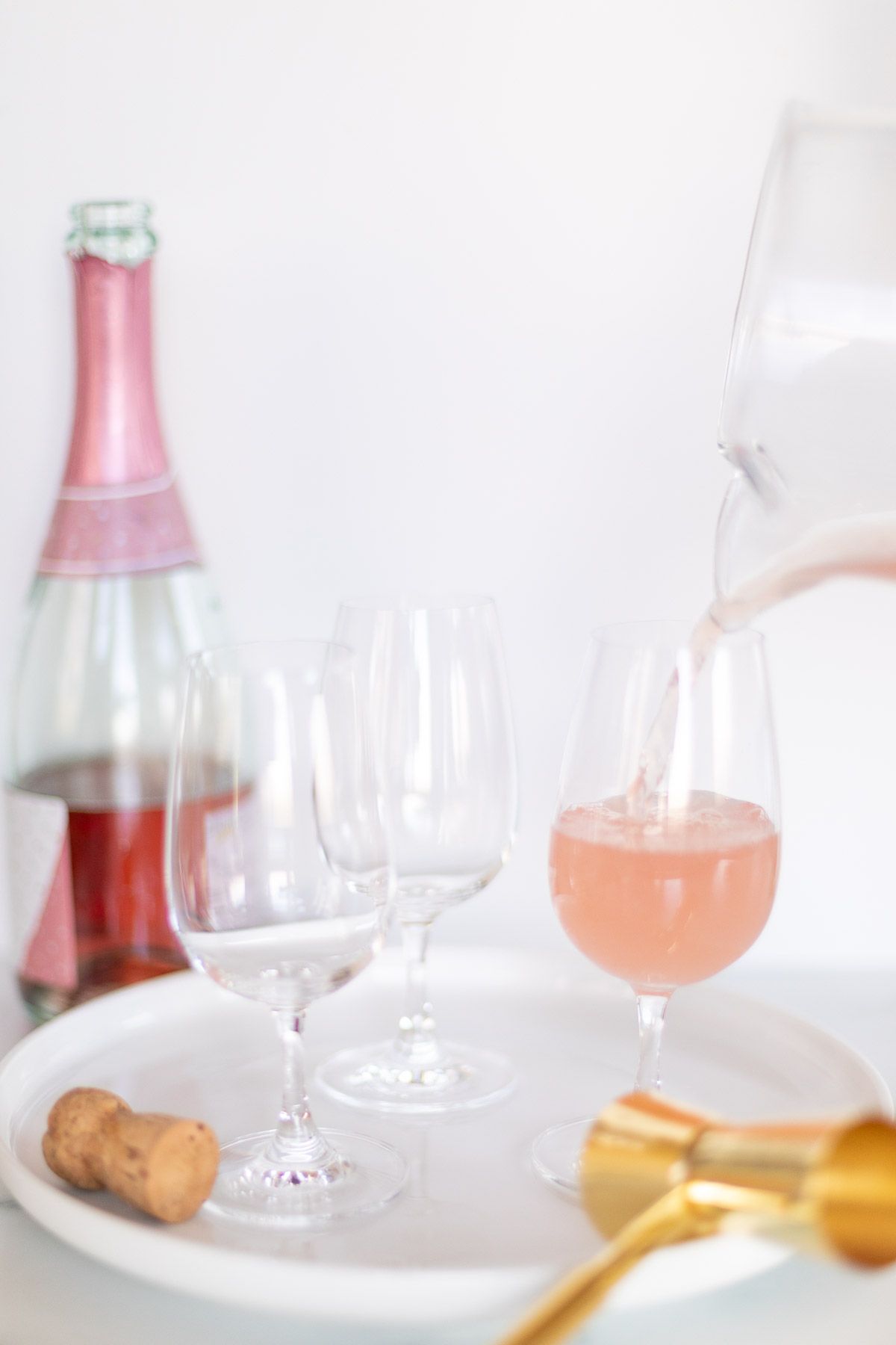 3 cocktail glasses on a white tray, with a glass pitcher pouring a pink cocktail into one.