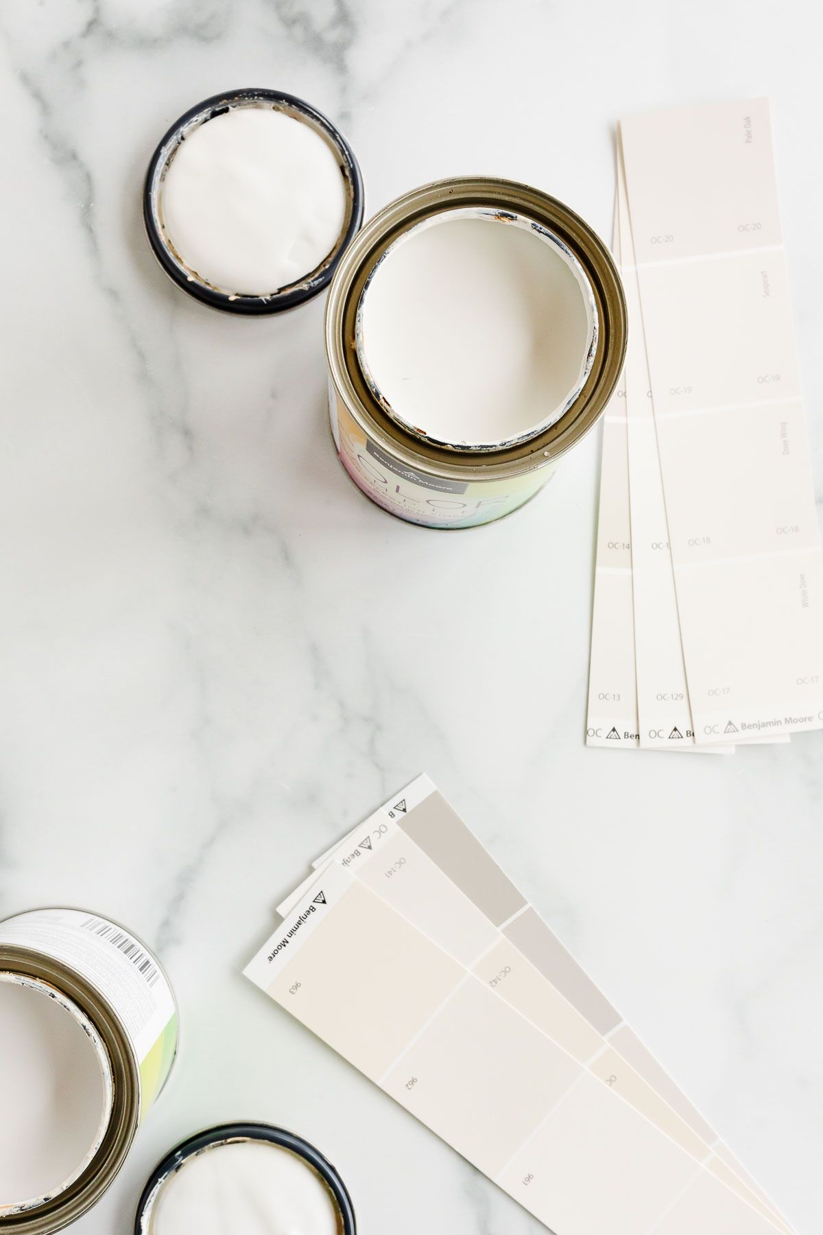 Open cans of warm whites and paint swatch cards laid out on a marble surface.