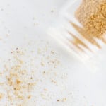 A glass jar tipped over on a white surface, spilling coarse brown beau monde seasoning.