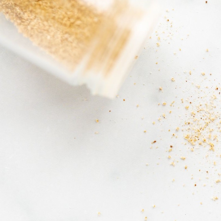 A clear glass spice jar of beau monde seasoning, spilled onto a white countertop