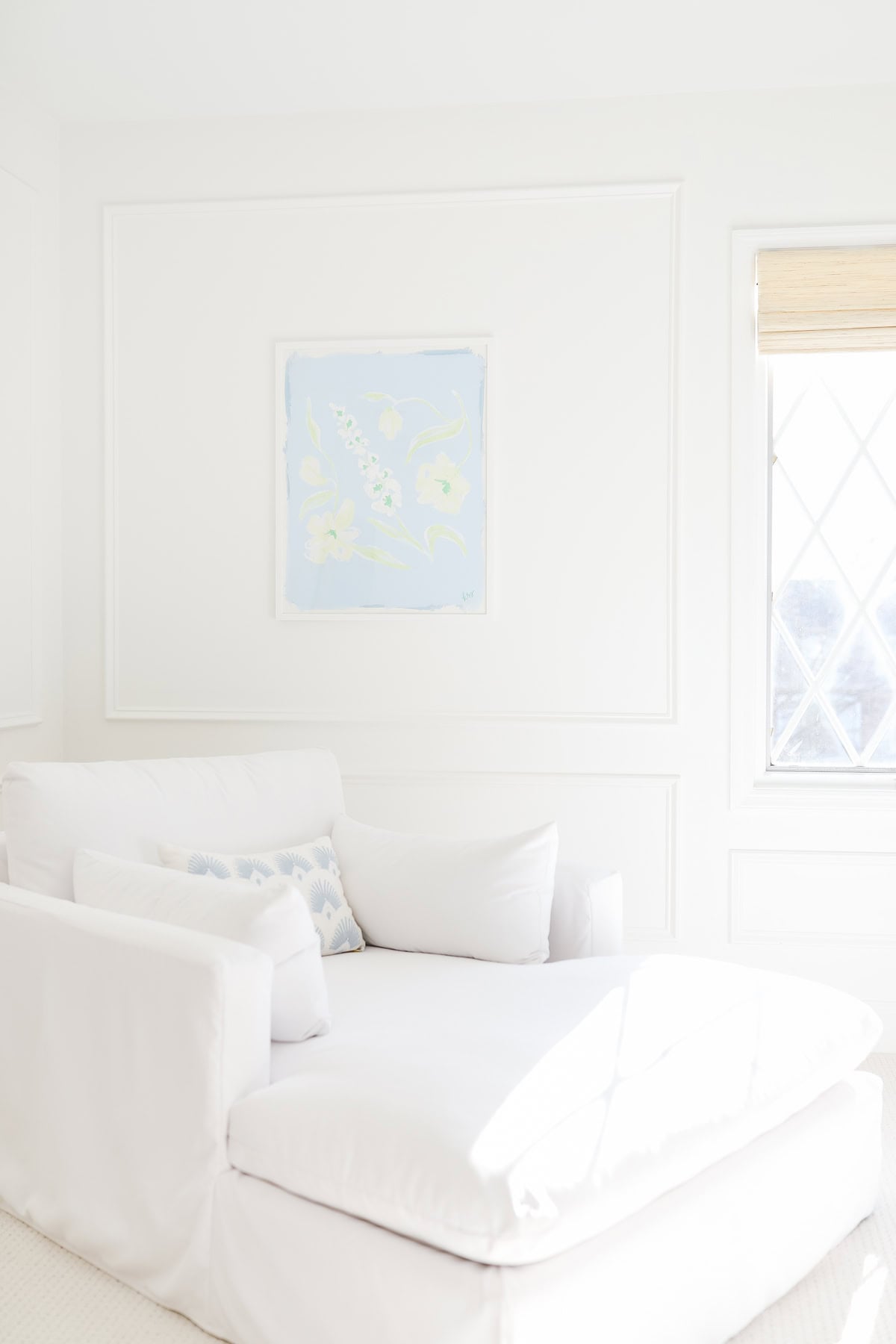 A white armchair with a patterned pillow sits in a bright room with white walls, romantic bedroom colors, a window with a blind, and light blue framed artwork on the wall.