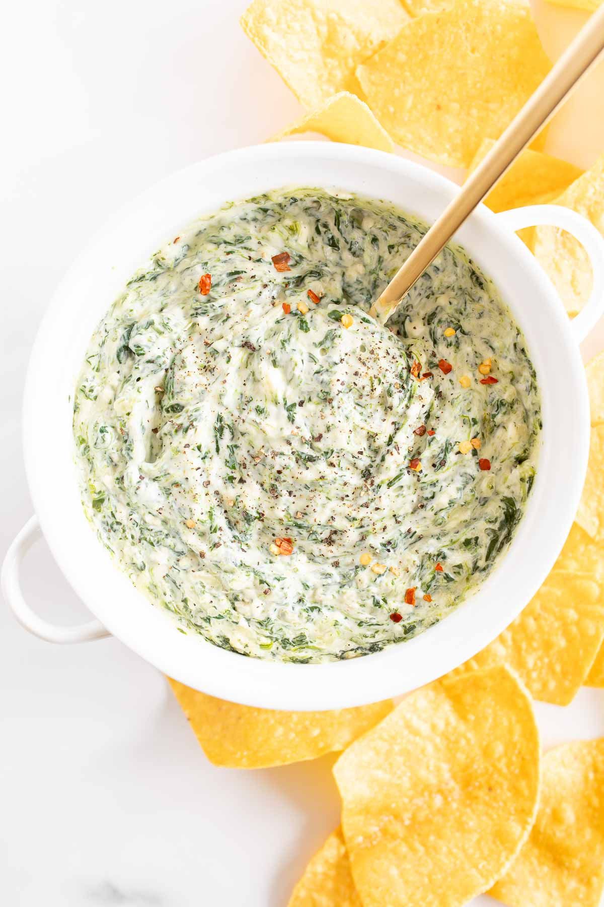 A white bowl full of spinach dip, surrounded by chips