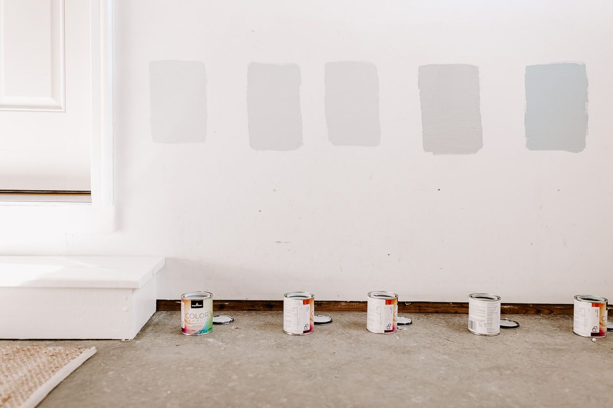 5 paint samples of blue gray paint colors on a white wall with the paint cans under them on the floor.