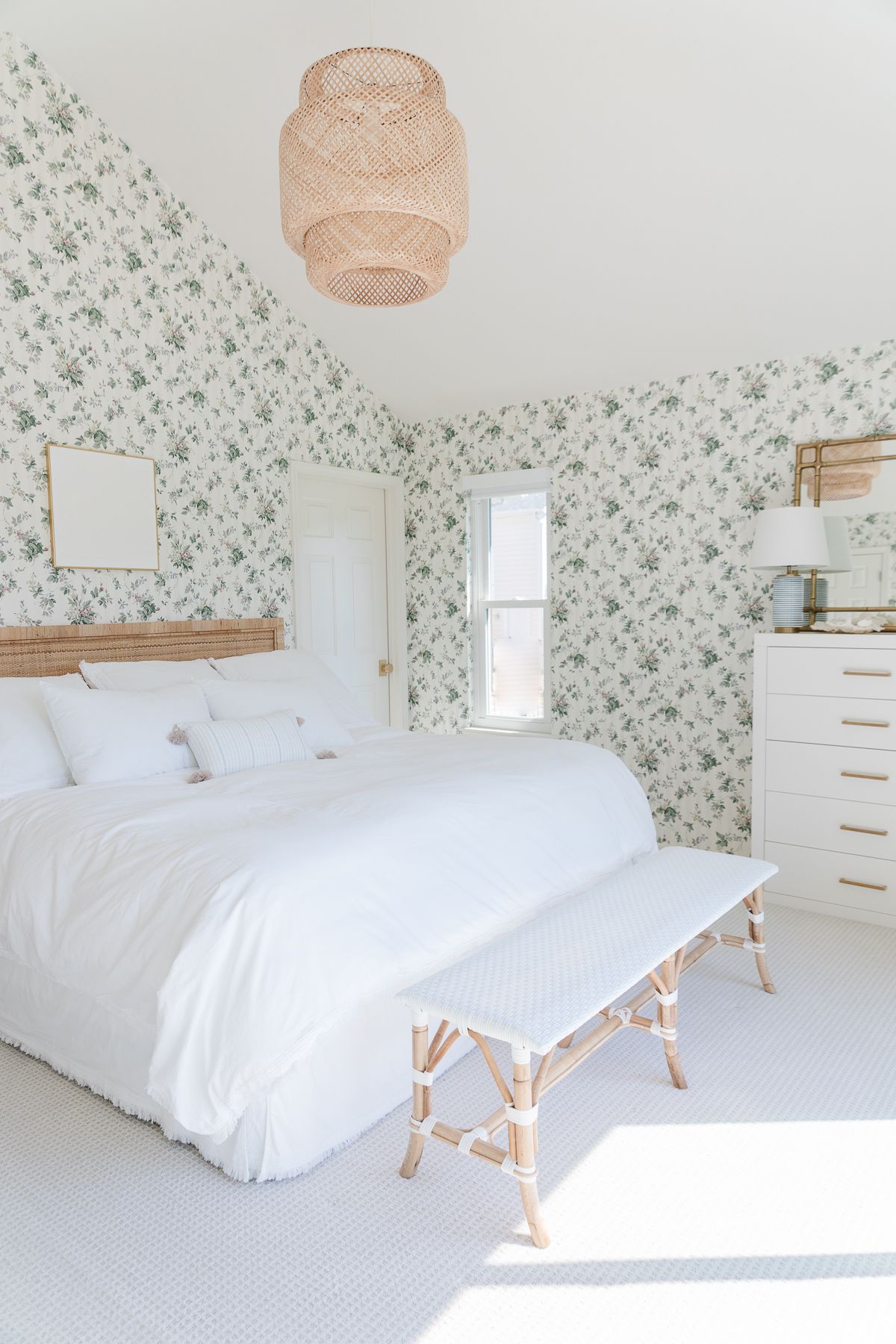 A coastal bedroom with floral wallpaper, and a rattan headboard, bench and light fixture