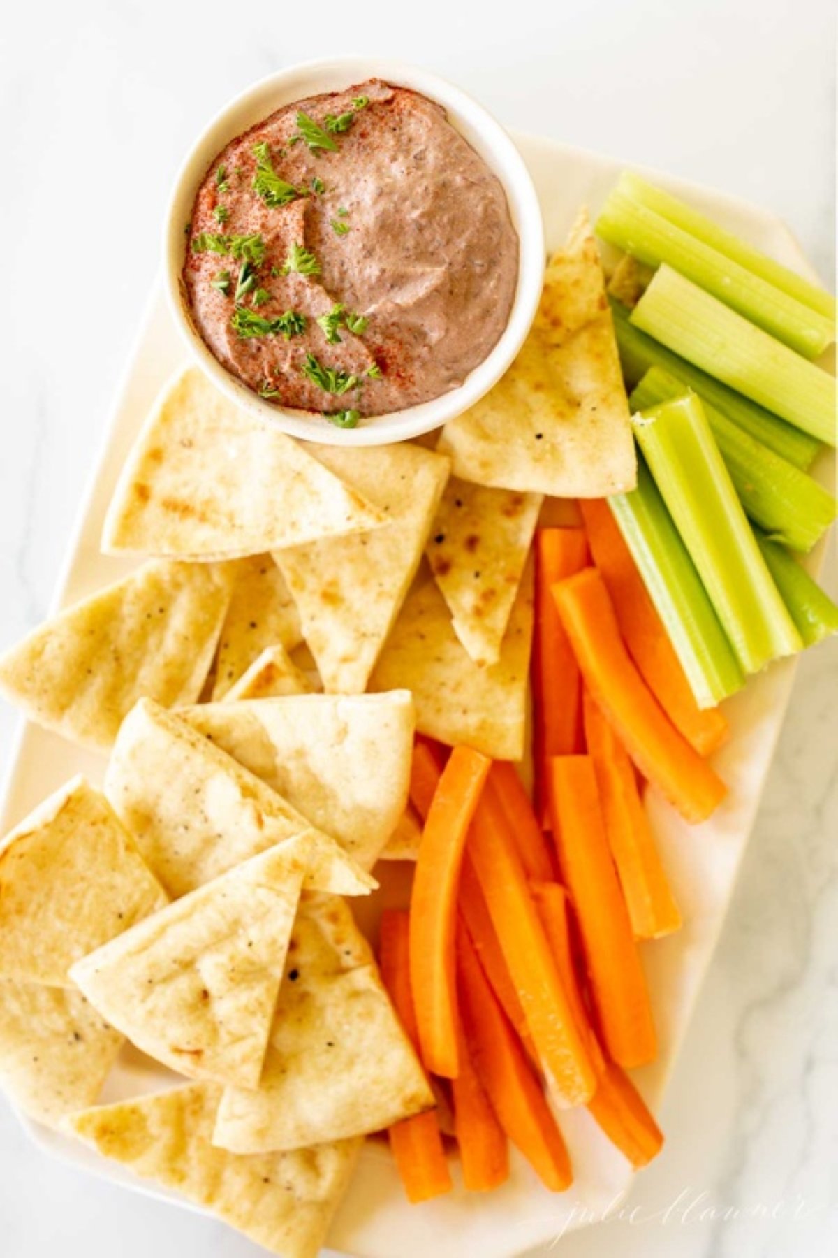 A white platter with a bowl of black bean hummus and pita triangles, celery and carrots.