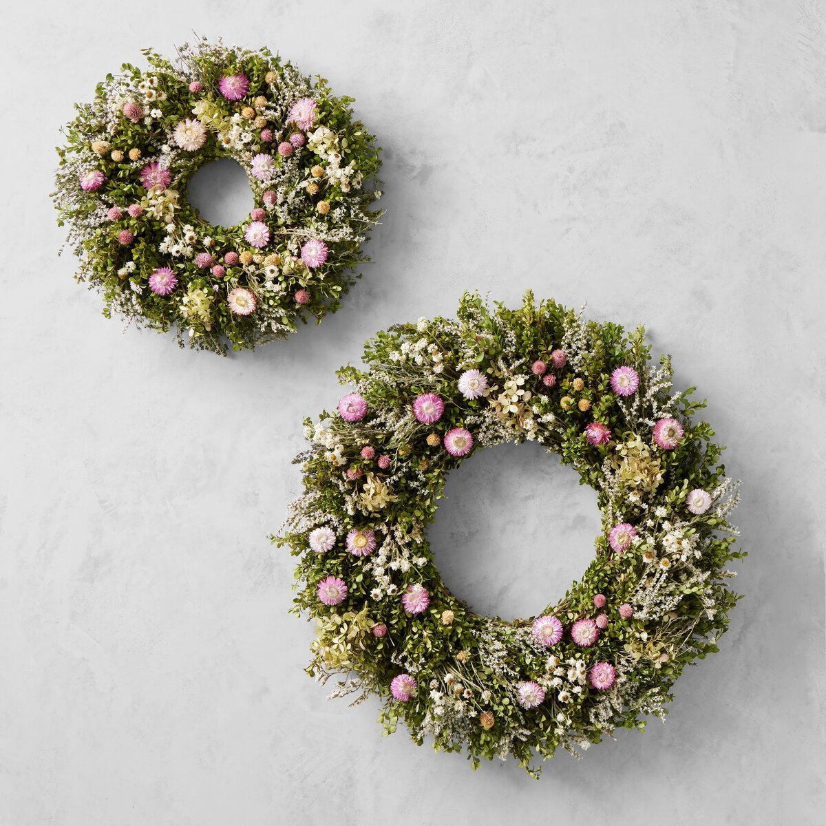 Two circular spring wreaths featuring pink, white, and green flowers with lush foliage are elegantly displayed against a light gray background.