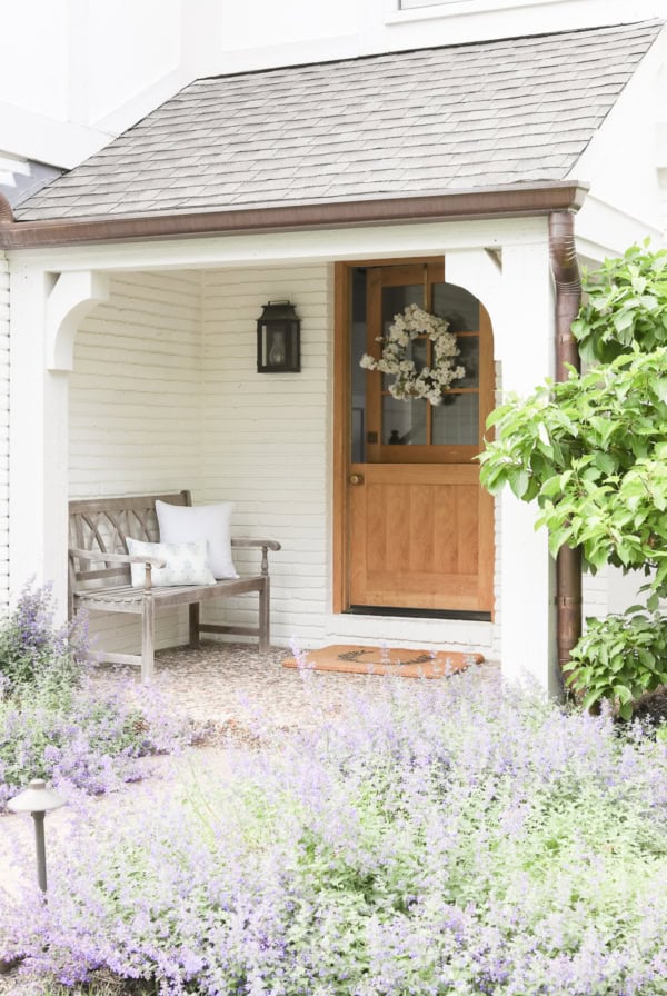 A small porch features a wooden door adorned with a spring wreath, a cozy bench with cushions, and a garden with purple flowers in the foreground.