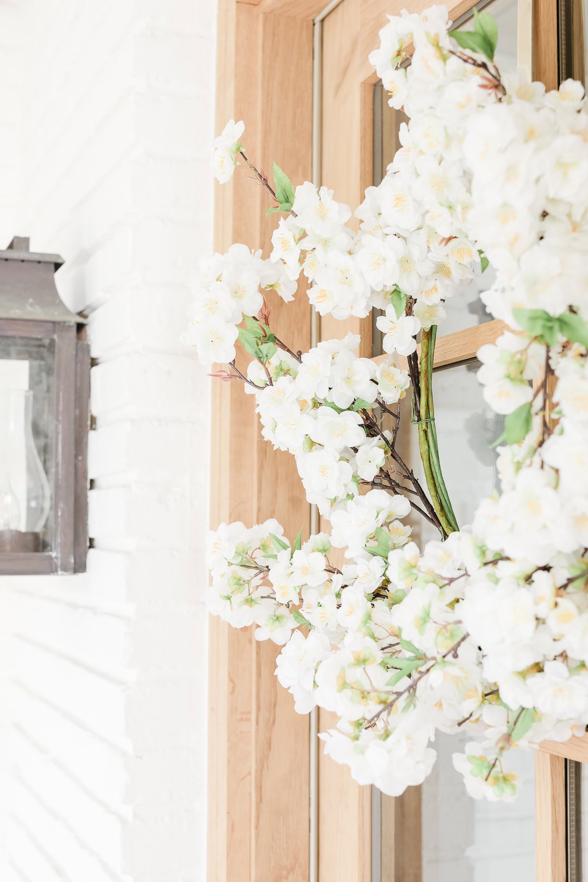 A spring wreath of white cherry blossoms graces a wooden door with a glass panel, complemented by a glass lantern sconce on the white brick wall.