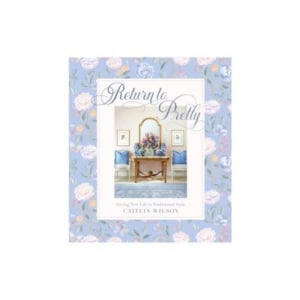 Book cover of "Return to Pretty" by Caitlin Wilson, featuring a floral border and a central image of a traditional styled room with a mirror, console table, two blue chairs, and inspiring nightstand decorating details.