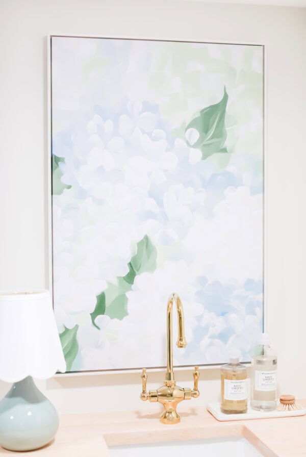 A gold faucet and sink beneath a pastel floral amazon art painting, with a lamp, soap dispenser, and bottle on a light wood countertop.