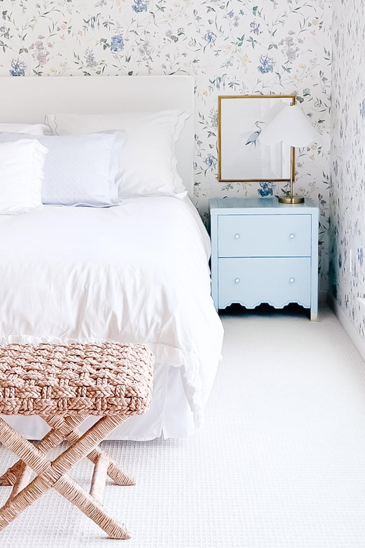 A neatly made bed with white linens sits beside a blue nightstand adorned with stylish nightstand decorating and a lamp; framed art rests beneath floral wallpapered walls.