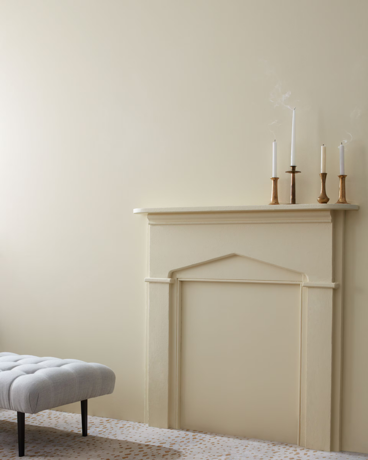 A beige fireplace mantel with three white candles in gold holders. Wall painted in Benjamin Moore Revere Pewter, is partially visible on the left side of the image. The floor has a light-colored, speckled pattern.