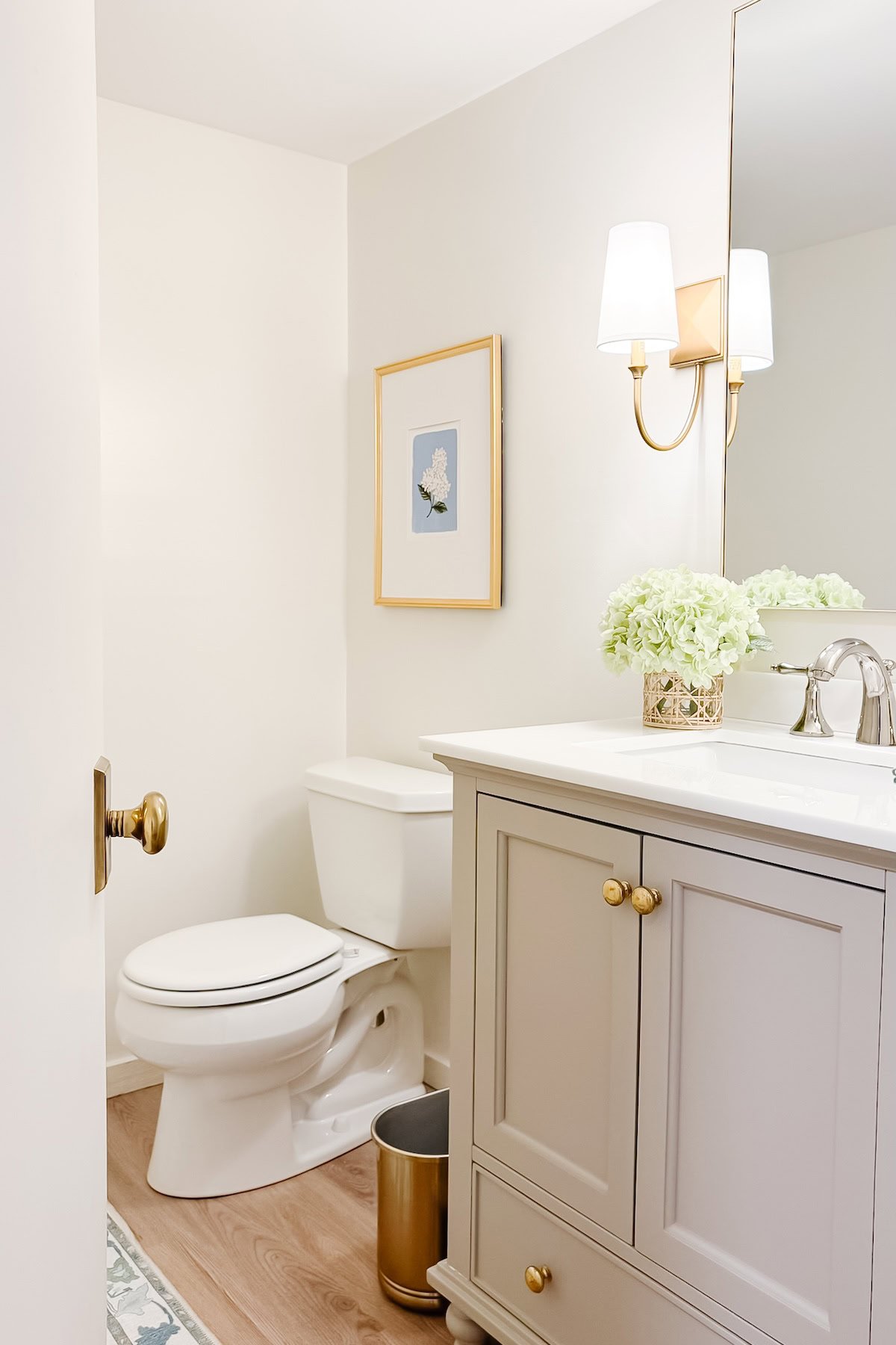 A bathroom with a white toilet, a gray vanity in Benjamin Moore Revere Pewter and brass hardware, a large mirror, wall sconce, framed art, flowers in a vase, and a small metal trash can on a wood floor.