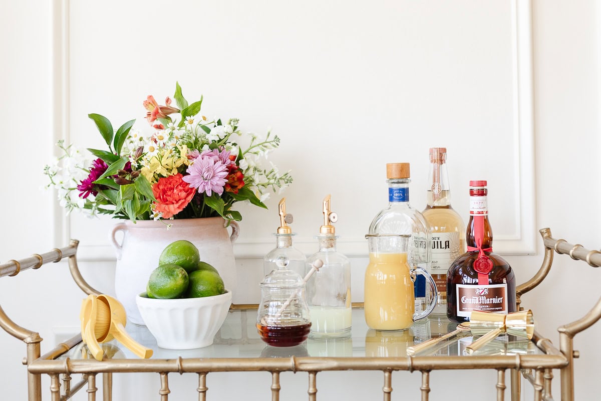 A vintage bar cart with an arrangement of colorful flowers in a vase, assorted liquor bottles, fresh limes, and a Cinco de Mayo menu, set against a white wall.