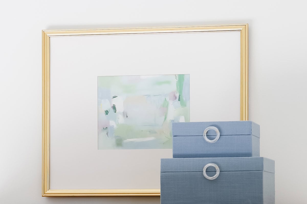 A gold-framed abstract amazon art painting hangs on a white wall above two stacked light blue storage boxes with circular handles.