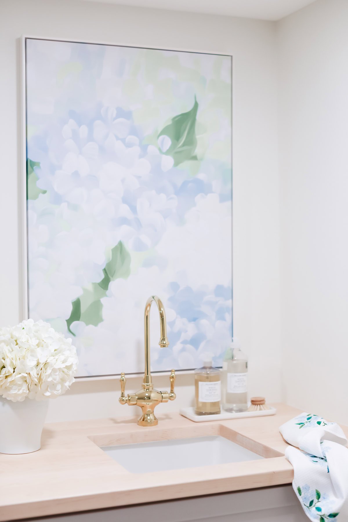 A small sink with a gold faucet, hand soap, and lotion bottles on a light wooden countertop, white hydrangeas in a vase, and a large amazon art floral painting on the wall above.